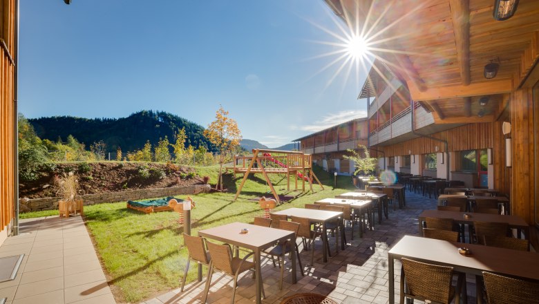 Terrasse des JUFA Hotels Annaberg mit Tischen, St&uuml;hlen und Spielplatz im Hintergrund bei Sonnenschein.
