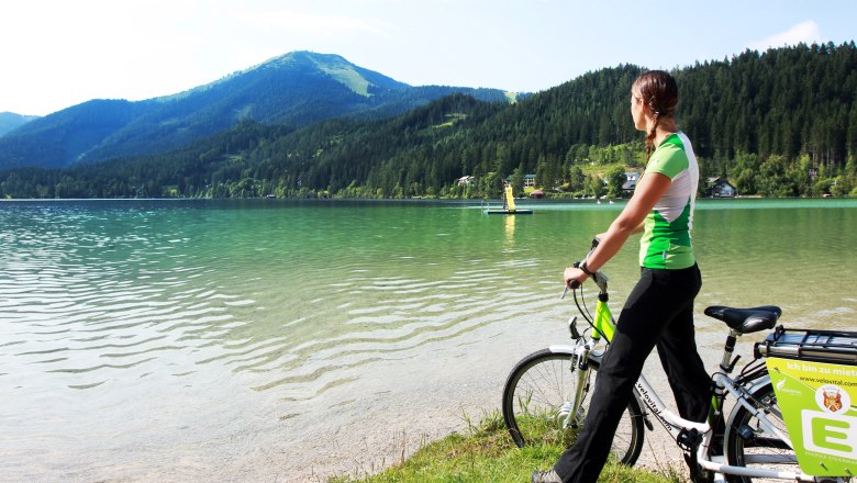 Frau mit Fahrrad am Ufer des Erlaufsees, umgeben von Bergen und Wald.