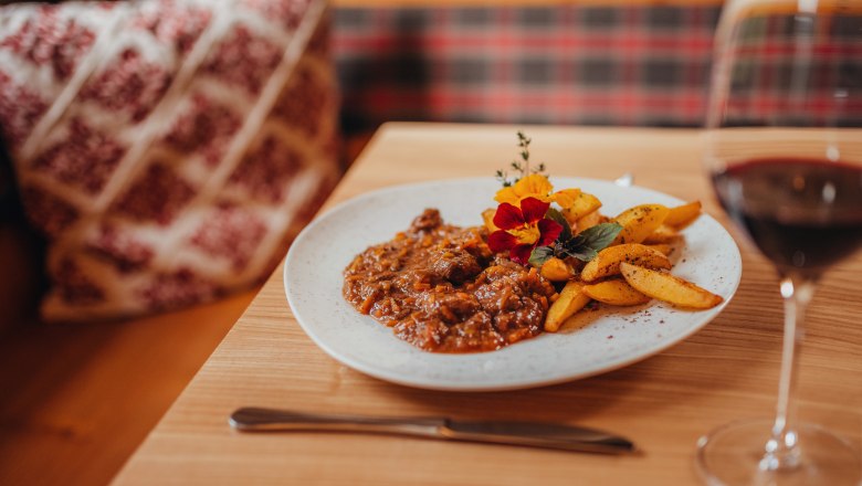 A plate of lamb ragout and thyme potatoes, decorated with edible flowers, on a wooden table. A glass of red wine stands next to it.