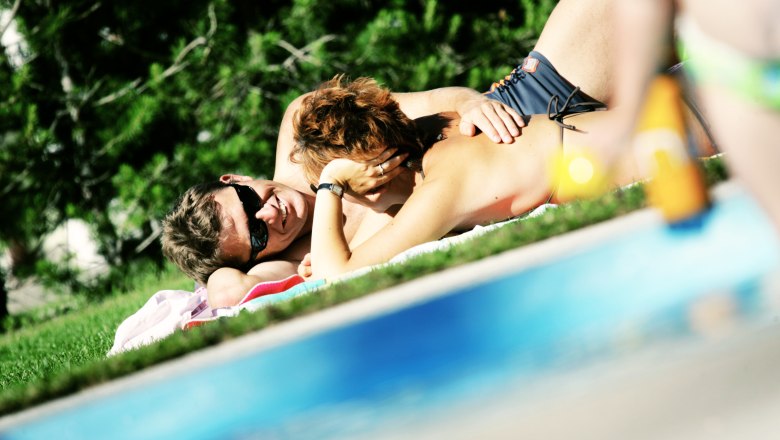 Ein Paar liegt entspannt auf einer Wiese neben einem Pool.