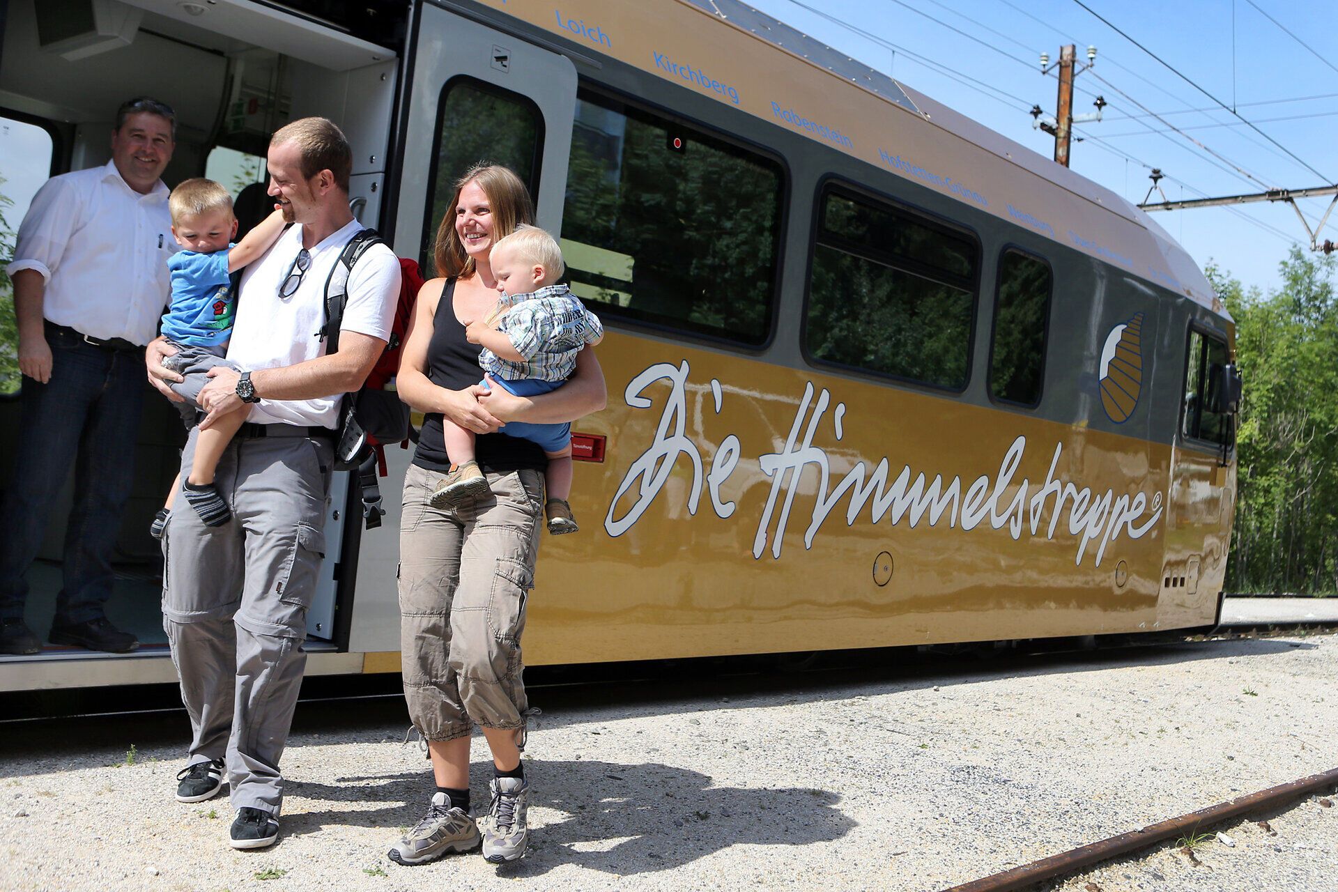 Eine fröhliche Familie steigt aus der bunten Bahn und genießt den warmen Sommertag. Die Kinder lachen und tragen ihre kleinen Rucksäcke, während die Eltern die malerische Umgebung bewundern. Hier beginnt ein unvergessliches Abenteuer in den Bergen.