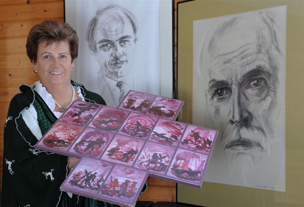 Frau in Tracht hält Kunstwerk vor zwei Porträts.