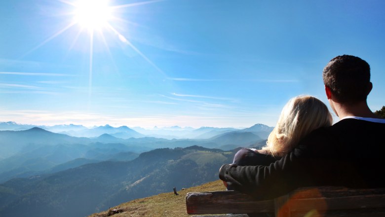 Ein Paar sitzt auf einer Bank und blickt auf eine Berglandschaft unter einem strahlend blauen Himmel mit Sonne.