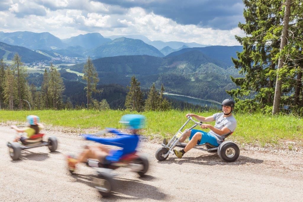 Personen fahren mit Mountaincarts den Berg hinunter, eine beeindruckende Berglandschaft im Hintergrund.