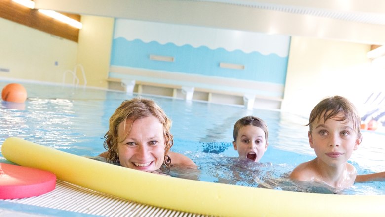 A woman and two children swim in an indoor pool, smiling and playing with a yellow pool noodle.