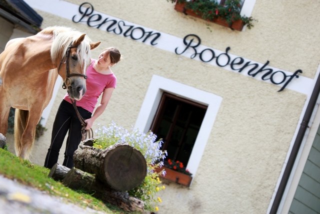 Eine Frau steht mit einem Pferd vor einem Geb&auml;ude mit der Aufschrift 'Pension Bodenhof'.