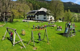 Kinder spielen auf einem Spielplatz vor einem gro&szlig;en Haus in einer gr&uuml;nen Landschaft.