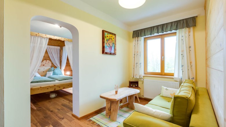 Gem&uuml;tliches Zimmer mit Holzm&ouml;beln, gr&uuml;nem Sofa, gro&szlig;em Fenster und einem Himmelbett im Hintergrund.