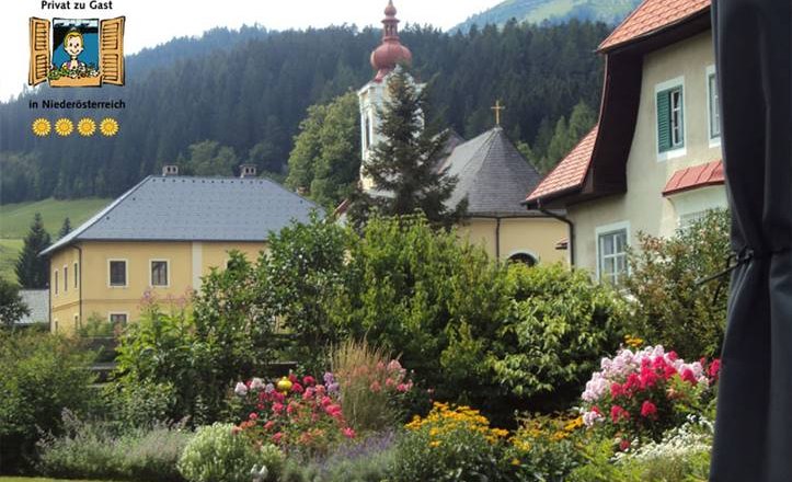 Haus Weissenbacher, © Haus Weissenbacher Ein malerischer Garten mit bunten Blumen vor traditionellen Häusern und einer Kirche in einer bergigen Landschaft.