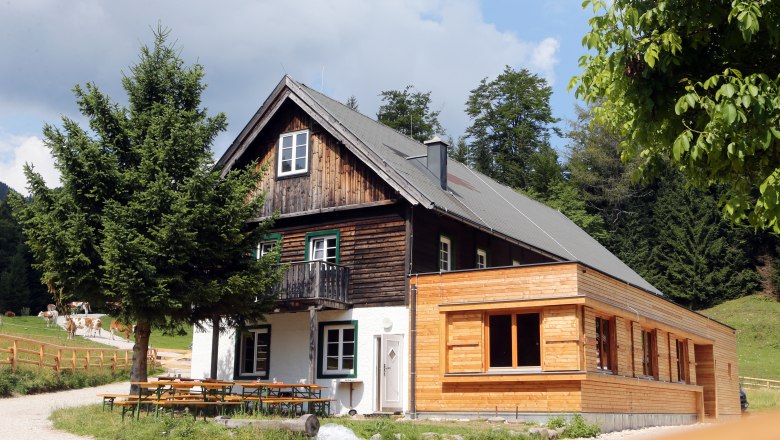Ein traditionelles Holzhaus mit Anbau in einer ländlichen Umgebung, umgeben von Bäumen und Wiesen.