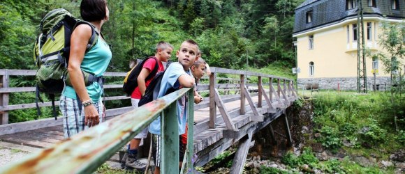 Eine Gruppe von Menschen steht auf einer Holzbrücke in einer grünen, bewaldeten Umgebung. Im Hintergrund ist ein gelbes Gebäude zu sehen.