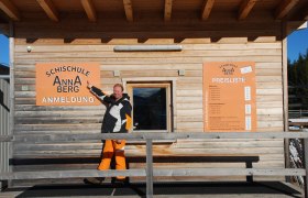 Ein Mann zeigt auf ein Schild der Schischule Annaberg vor einem Holzgebäude.