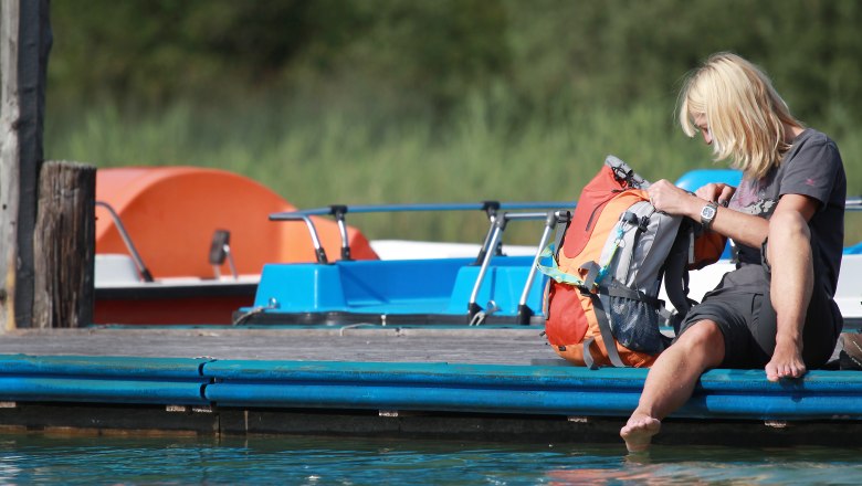 Person sitzt auf einem Steg am Erlaufsee mit den Füßen im Wasser, neben einem Rucksack. Im Hintergrund sind Tretboote zu sehen.