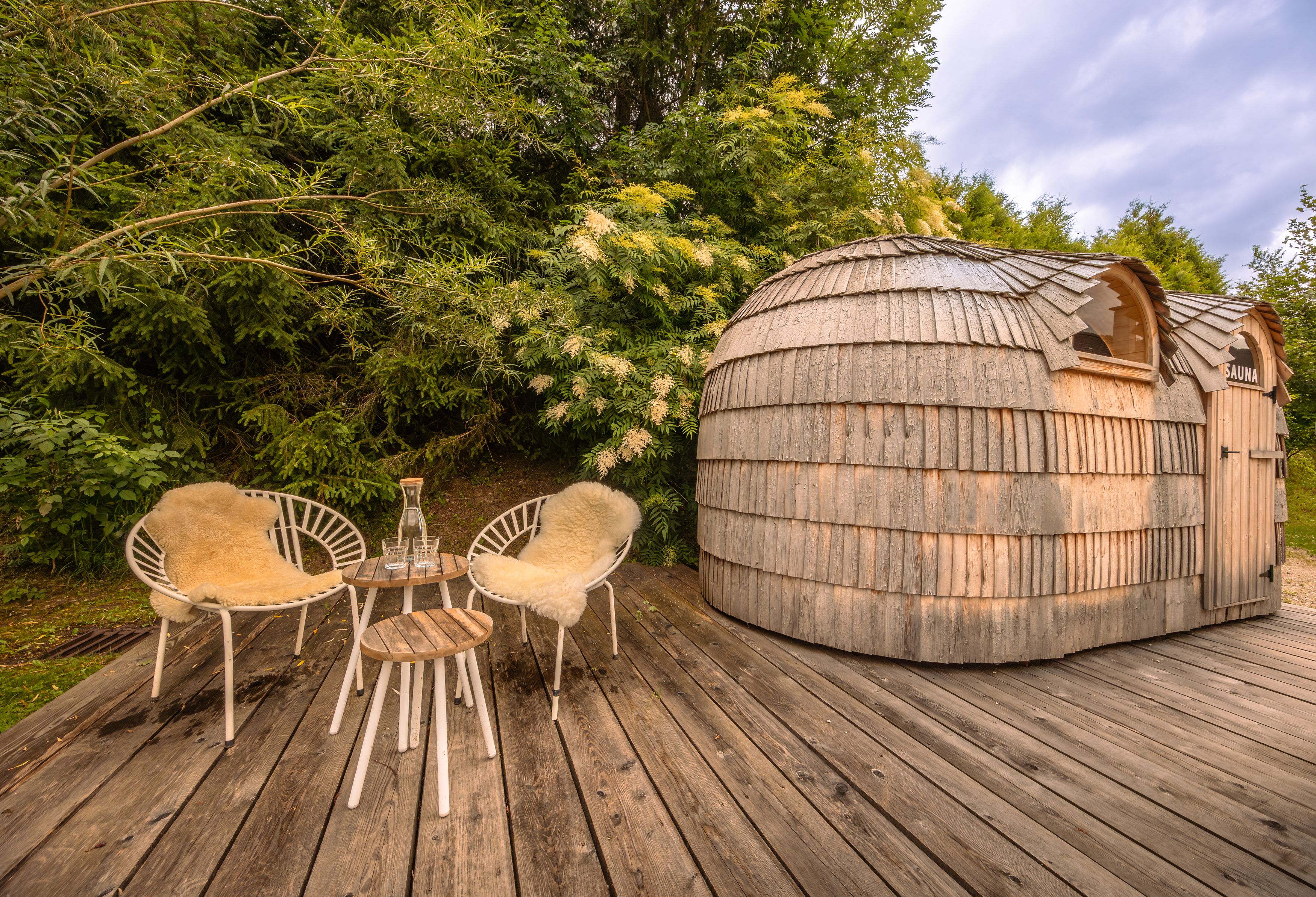 Wooden sauna with two chairs and table on a wooden terrace surrounded by greenery.