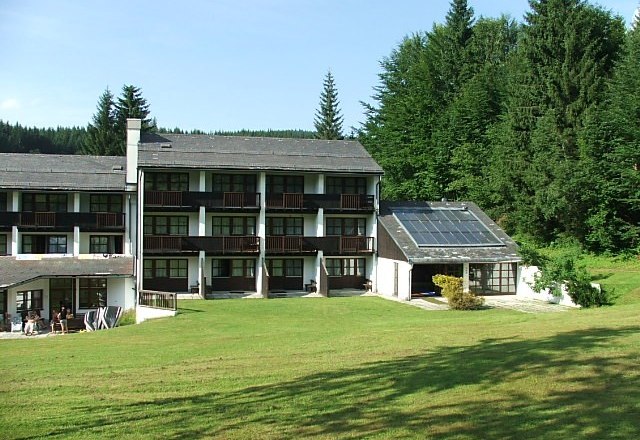Hotelgebäude mit Solarpanelen und grüner Wiese im Vordergrund, umgeben von Wald.