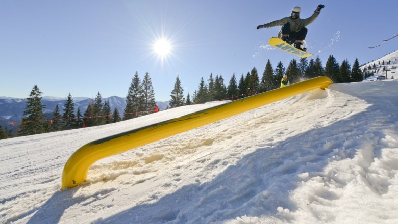Snowboarder springt über ein gelbes Hindernis auf einer verschneiten Piste bei Sonnenschein.