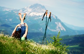 Person liegt auf einer Wiese mit Blick auf die Berge, Wanderst&ouml;cke daneben.