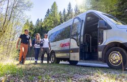 Drei wandernde Personen nähern sich einem Kleinbus, welcher bereits die Tür geöffnet hat. 