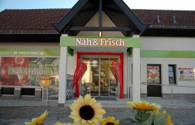 Eingang eines Nah & Frisch Supermarkts mit Sonnenblumen im Vordergrund.