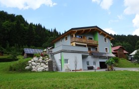 Ein modernes Haus mit Holzverkleidung und Solarpanelen in einer gr&uuml;nen, bewaldeten Umgebung.