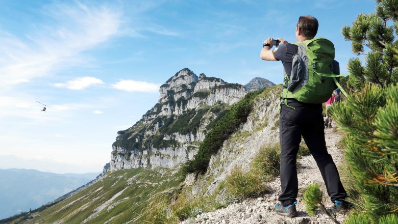 Ein Wanderer mit grünem Rucksack fotografiert eine Berglandschaft mit einem Hubschrauber am Himmel.