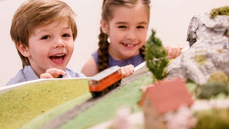 Zwei fr&ouml;hliche Kinder beobachten gebannt die vorbeifahrende Modelleisenbahn, w&auml;hrend sie in eine Welt voller Abenteuer und Fantasie eintauchen. Die detailreiche Landschaft mit kleinen H&uuml;tten und B&auml;umen l&auml;sst die Herzen der kleinen Eisenbahnfans h&ouml;her schlagen. Ein unvergesslicher Moment voller Staunen und Freude.