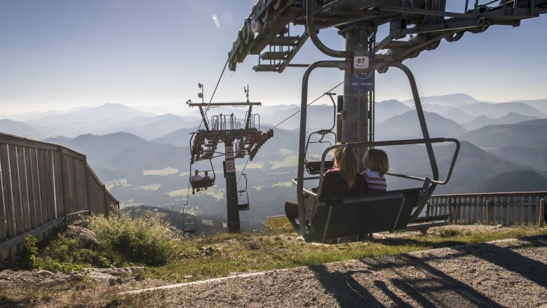 Two people on a chair lift with a mountain landscape in the background.