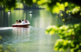 Vier Personen in einem Boot auf einem ruhigen See, umgeben von gr&uuml;ner Natur.